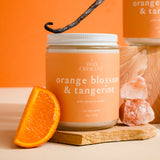 Candle jar labeled 'Orange Blossom & Tangerine' with an orange slice and vanilla bean on a wooden board against an orange background.