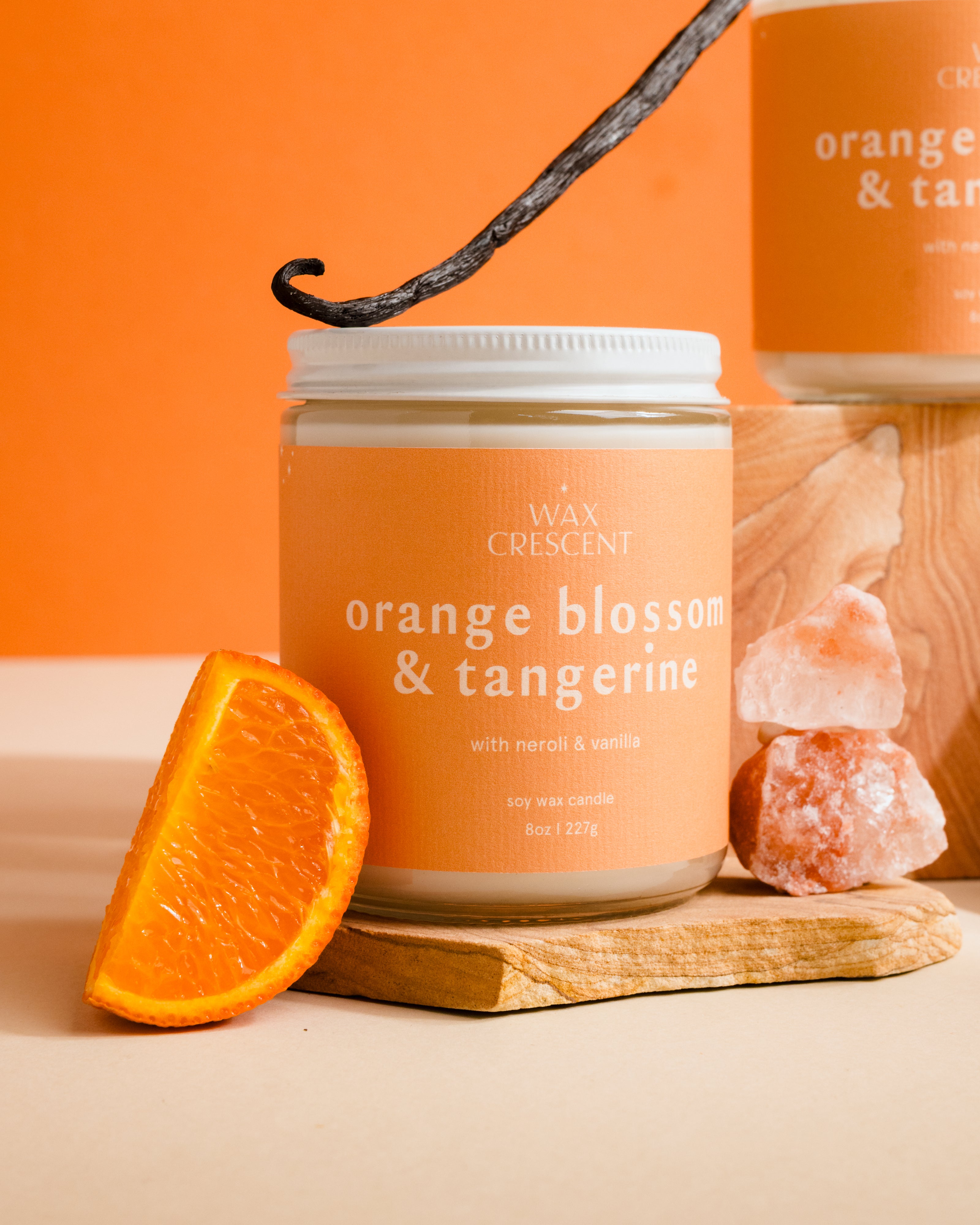 Candle jar labeled 'Orange Blossom & Tangerine' with an orange slice and vanilla bean on a wooden board against an orange background.