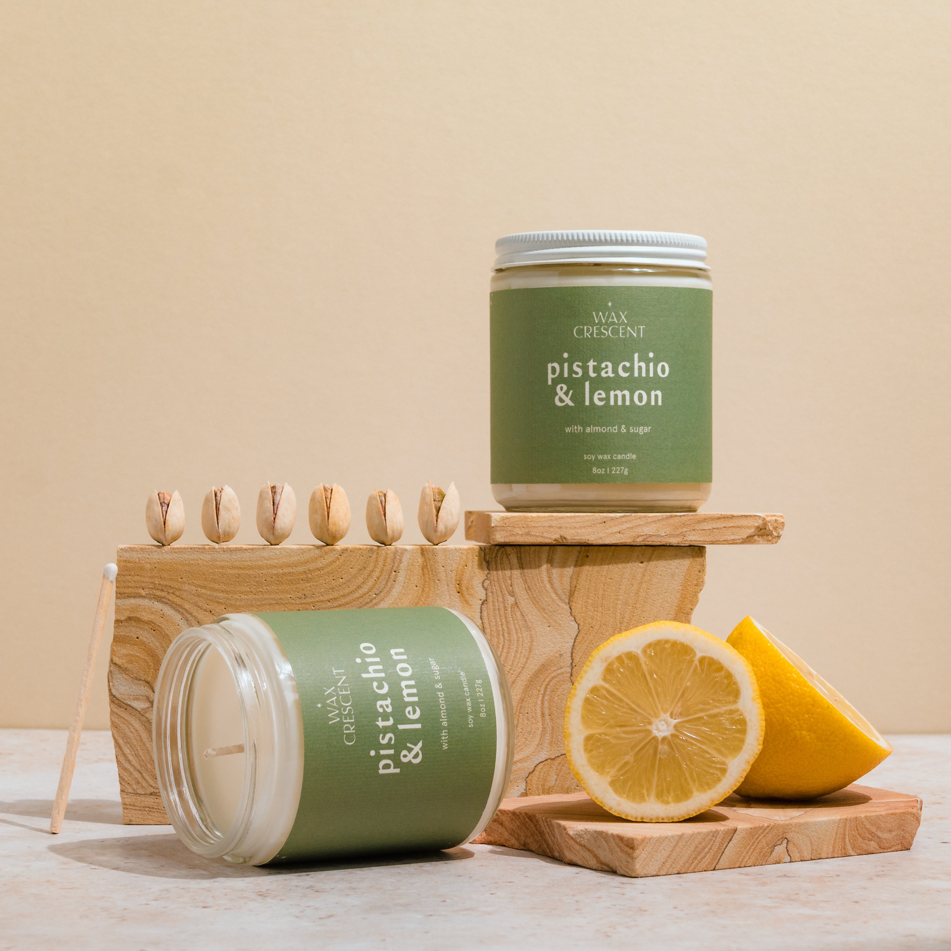 Two Pistachio & Lemon soy wax candles, one lying on its side, surrounded by fresh lemons and pistachios. The scene highlights the natural, refreshing ingredients that inspire the candle's fragrance.