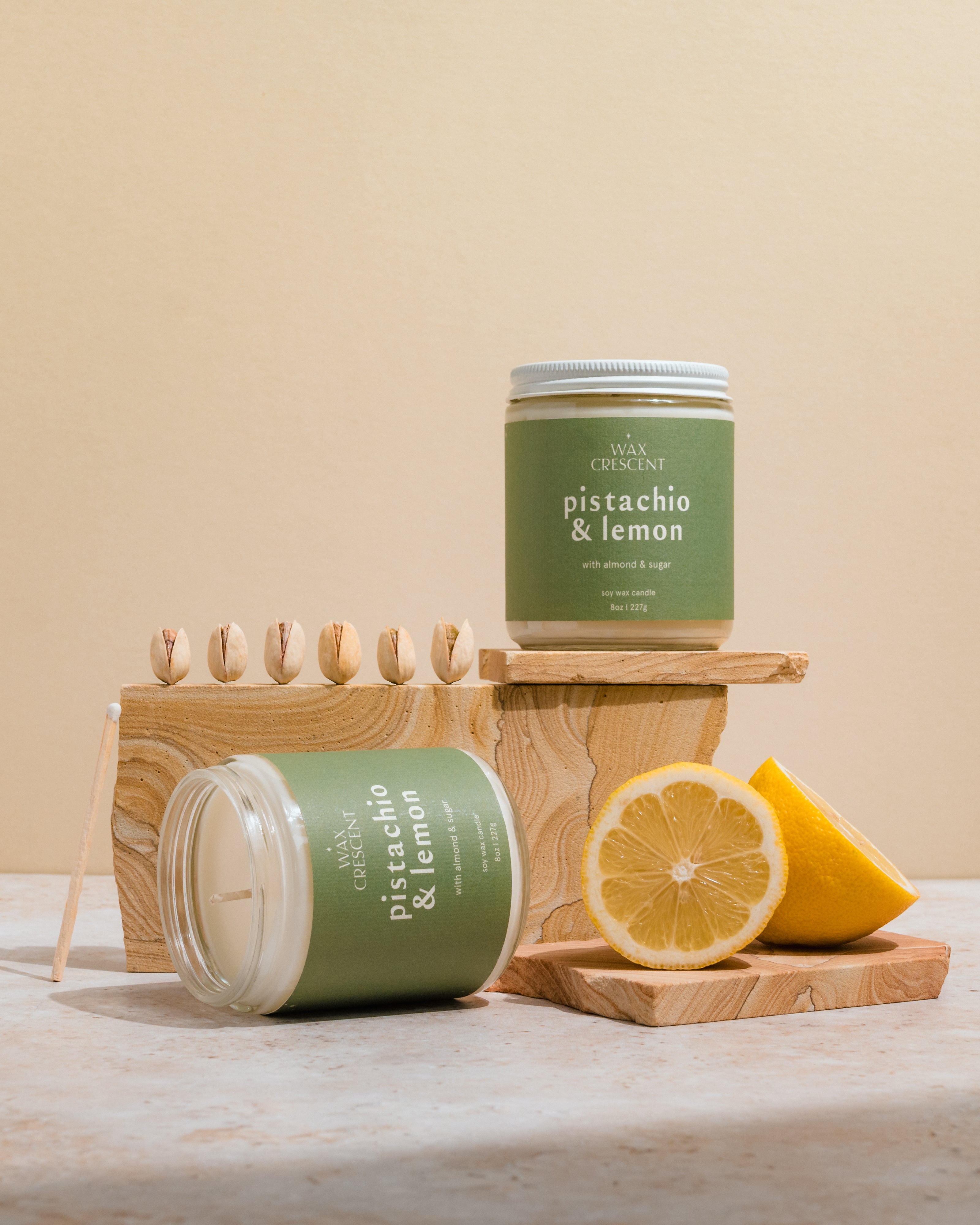 Two Pistachio & Lemon soy wax candles, one lying on its side, surrounded by fresh lemons and pistachios. The scene highlights the natural, refreshing ingredients that inspire the candle's fragrance.