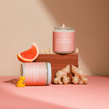 Pink candle with ginger and cardamom packaging on a pink surface with ginger slices and cardamom pods.