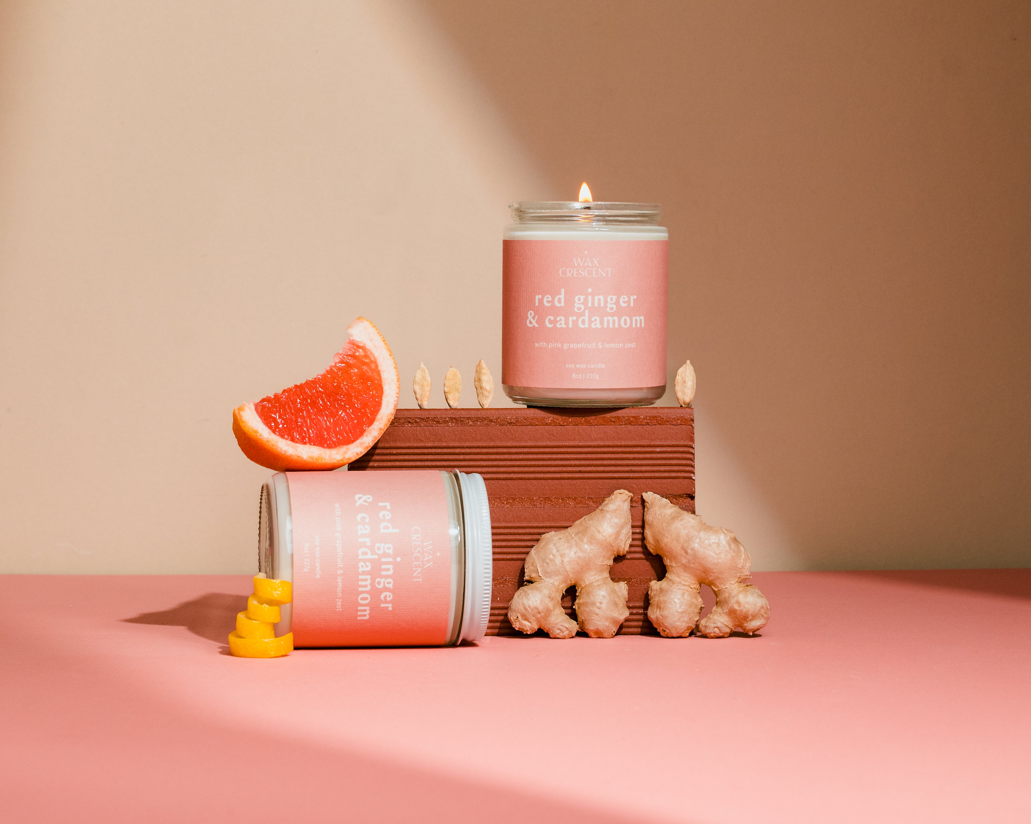 Pink candle with ginger and cardamom packaging on a pink surface with ginger slices and cardamom pods.