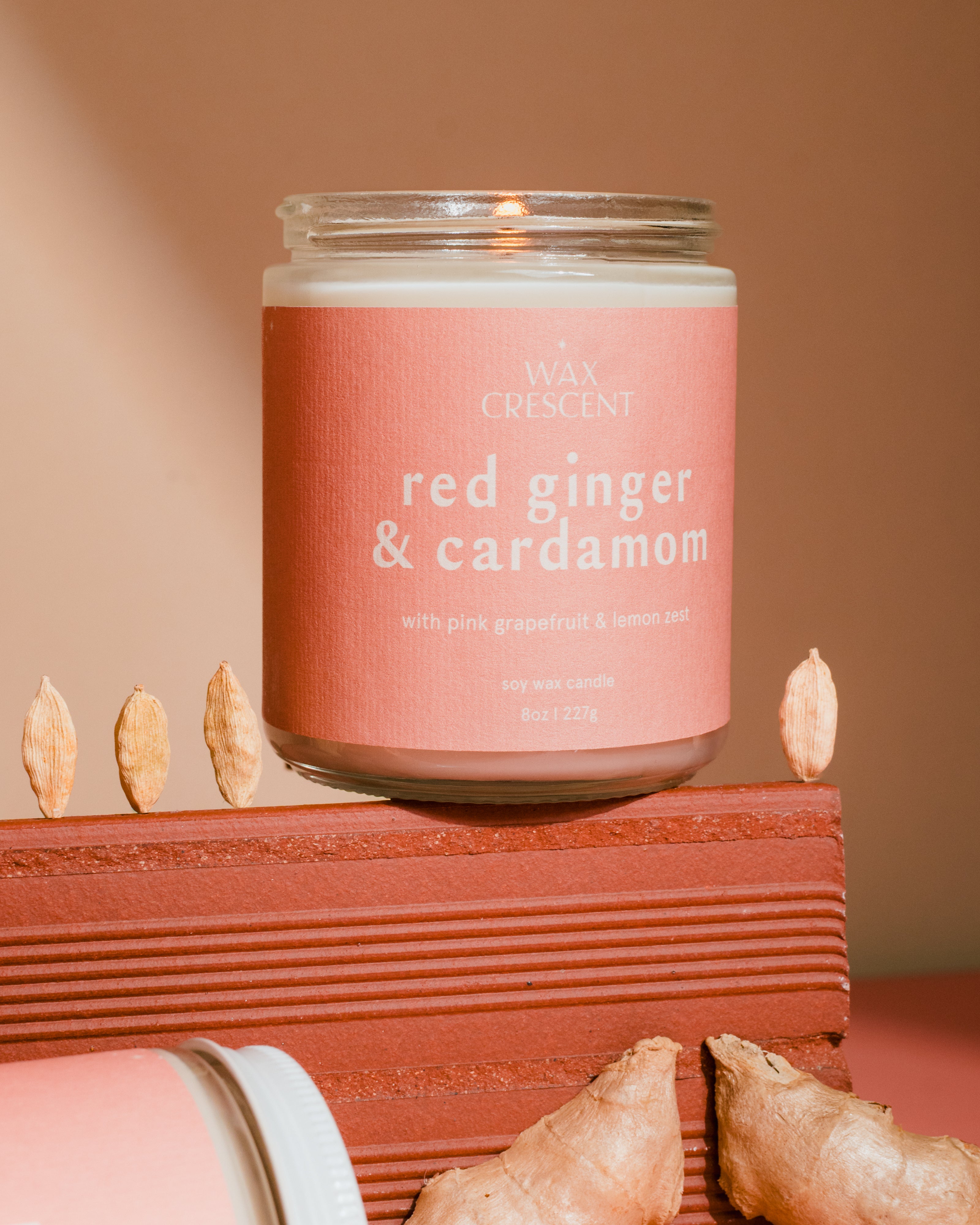 Wax Crescent candle labeled 'red ginger & cardamom' on a wooden block with ginger pieces.