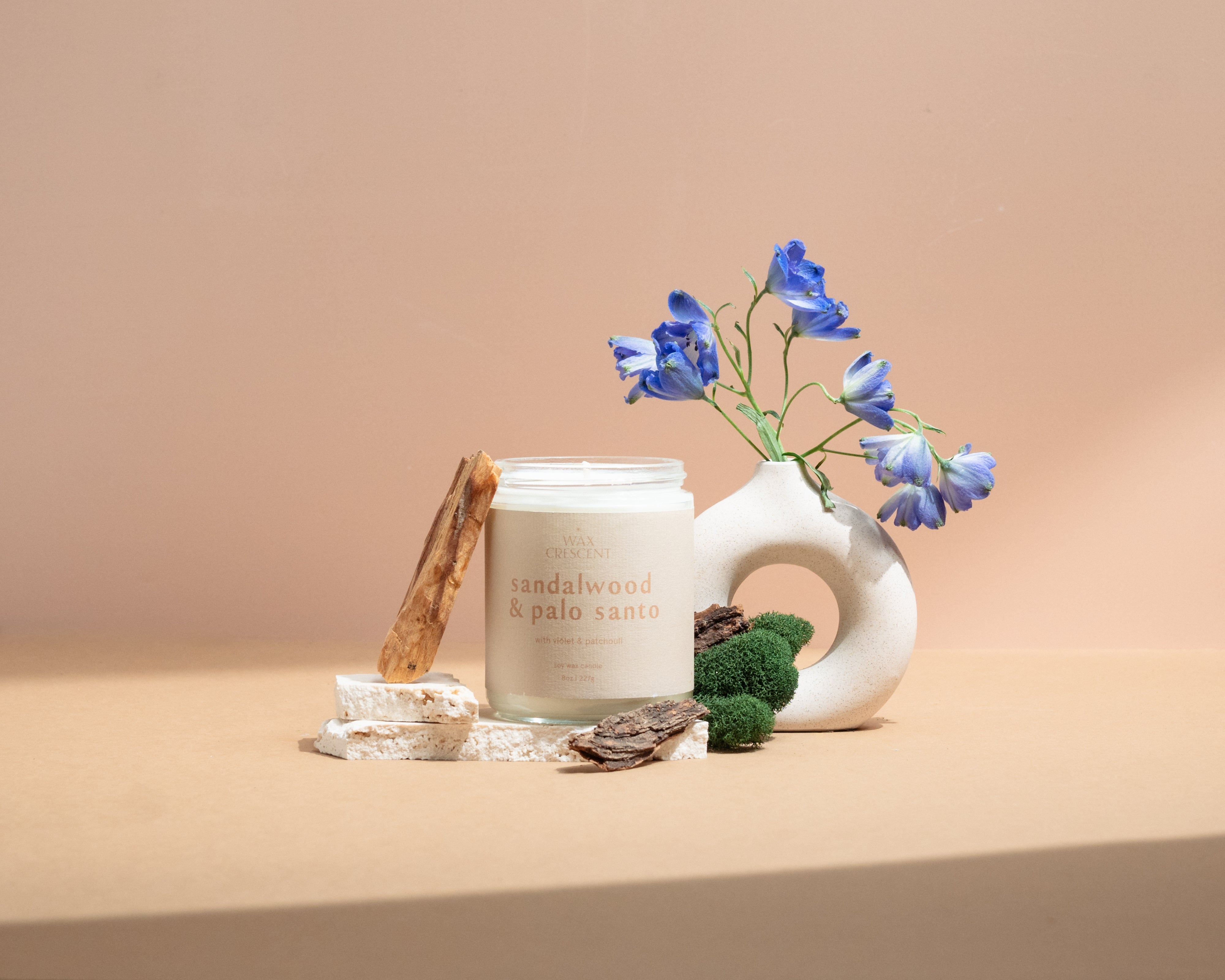 Wax Crescent Sandalwood & Palo Santo luxury soy candle surrounded by natural decor, including palo santo wood, moss, bark, and blue delphinium flowers, styled against a minimalist beige background. Perfect for creating a calming and meditative ambiance