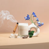 Wax Crescent Sandalwood & Palo Santo luxury soy candle surrounded by natural decor, including palo santo wood, moss, bark, and blue delphinium flowers, styled against a minimalist beige background. Perfect for creating a calming and meditative ambiance