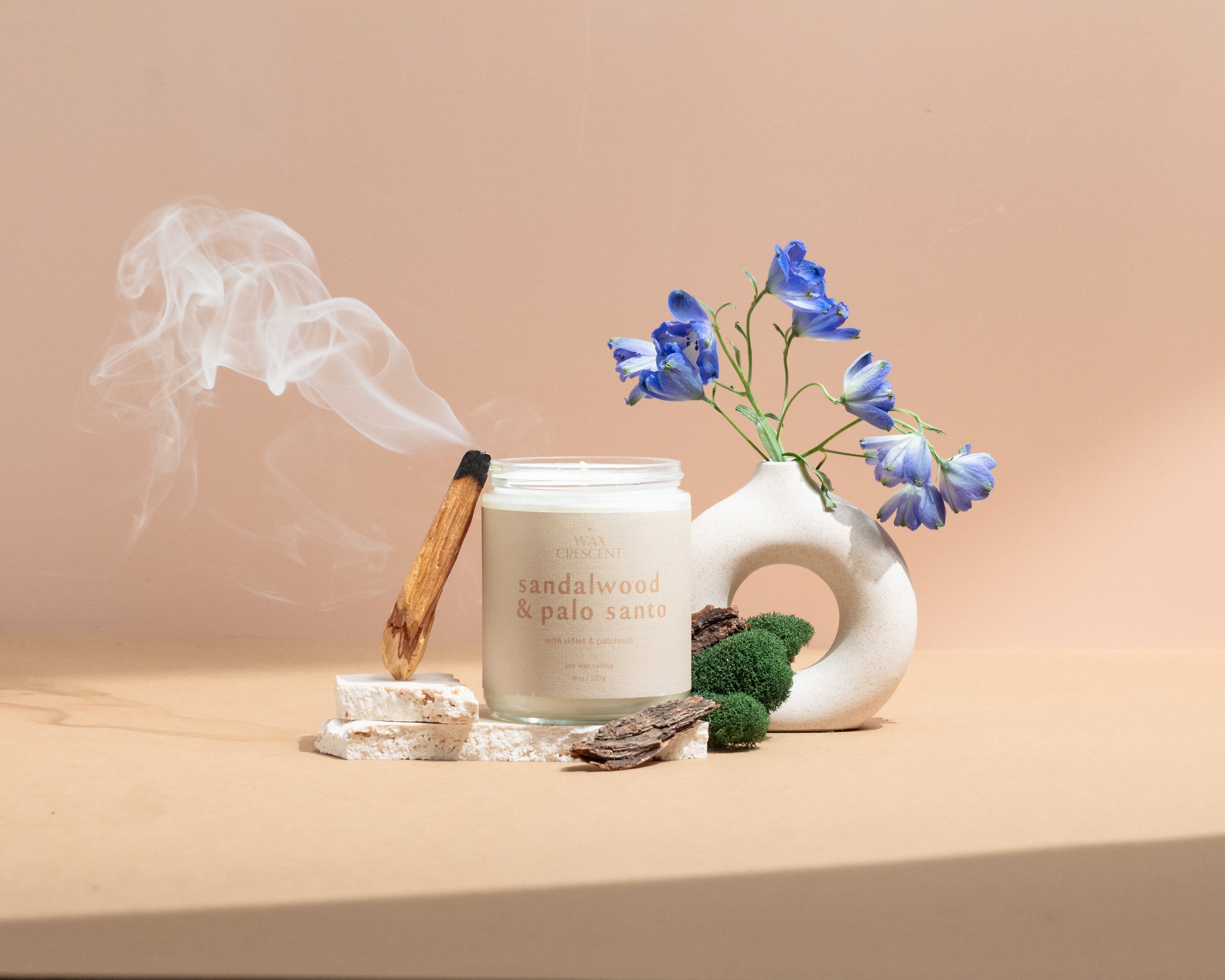 Wax Crescent Sandalwood & Palo Santo luxury soy candle surrounded by natural decor, including palo santo wood, moss, bark, and blue delphinium flowers, styled against a minimalist beige background. Perfect for creating a calming and meditative ambiance