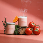Wax Crescent Tomato & Vetiver candle surrounded by ripe tomatoes and orange citrus on a pink backdrop, inspired by fresh summer gardens.