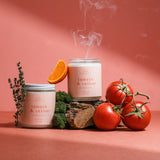 Wax Crescent Tomato & Vetiver candle surrounded by ripe tomatoes and orange citrus on a pink backdrop, inspired by fresh summer gardens.