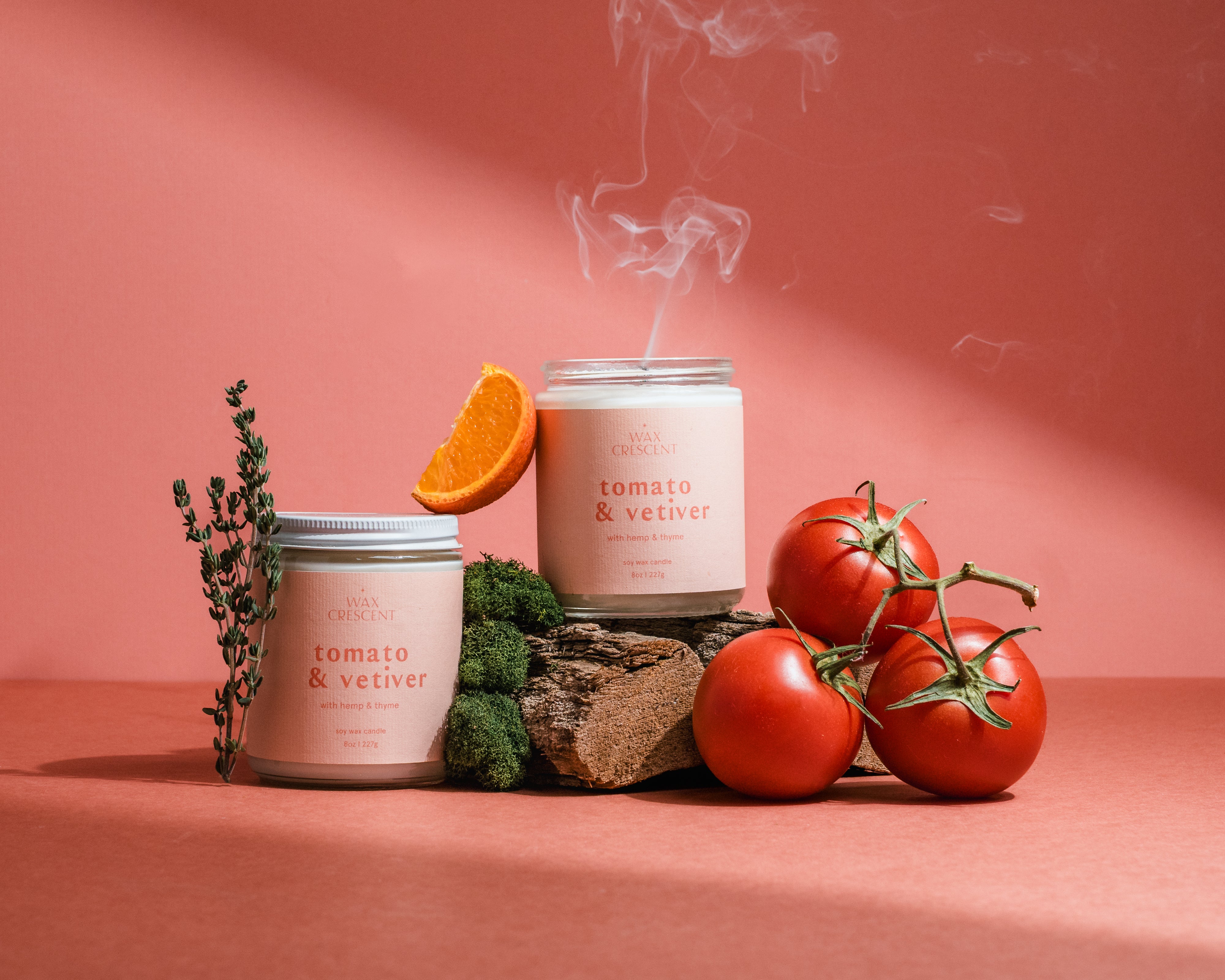 Wax Crescent Tomato & Vetiver candle surrounded by ripe tomatoes and orange citrus on a pink backdrop, inspired by fresh summer gardens.