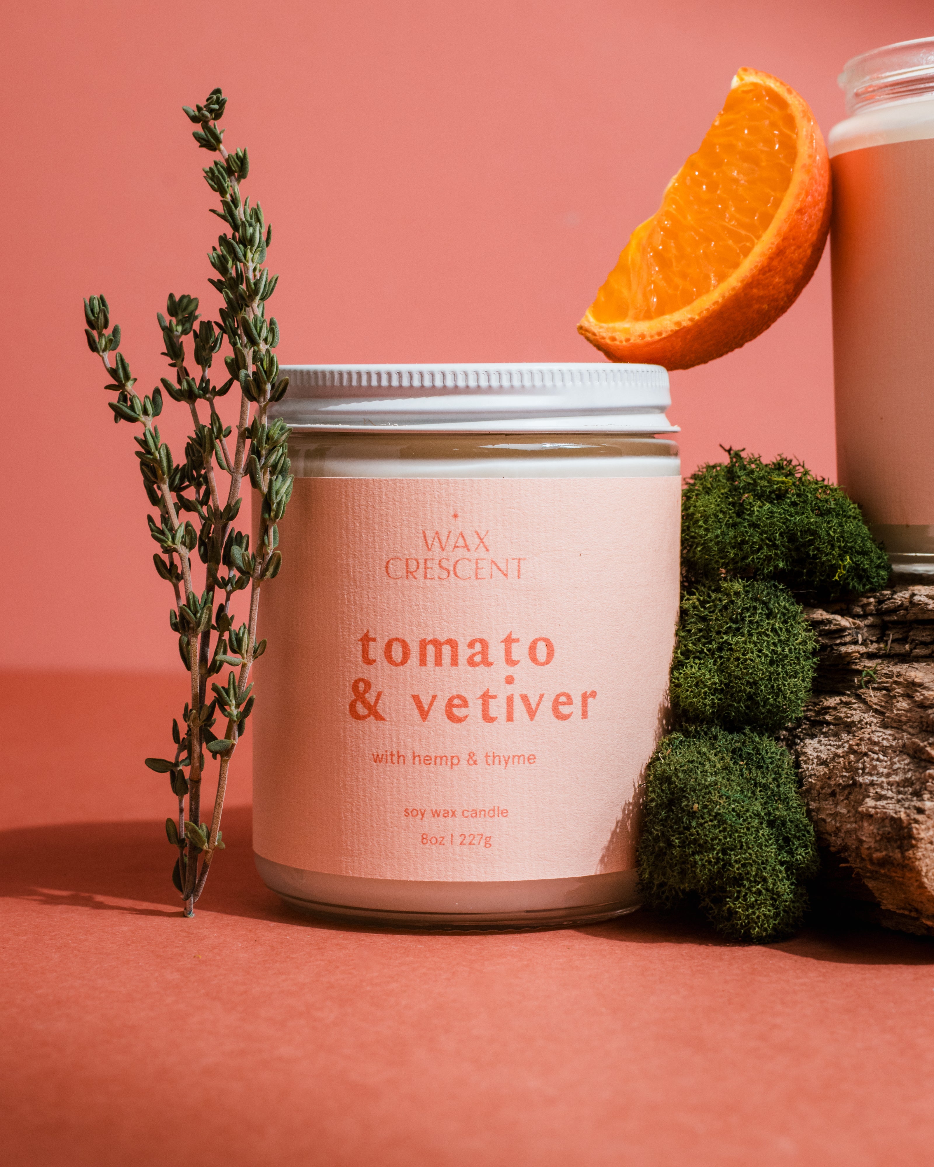 Clean-burning Tomato & Vetiver candle by Wax Crescent styled with fresh tomatoes and orange on a pink background for elevated summer home fragrance.