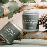 Two candles labeled 'White Birch & Eucalyptus' on a branch with leaves and a pine cone.