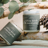 Two candles labeled 'White Birch & Eucalyptus' on a branch with leaves and a pine cone.