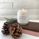 Lit Cranberry & Balsam candle on a leather book with pine cones, creating a cozy, rustic ambiance with a fresh, balsam scent