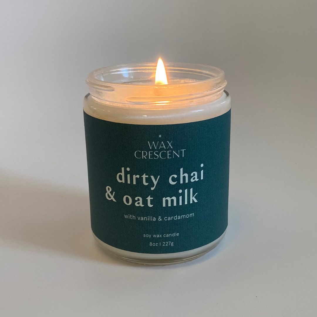 Wax Crescent Dirty Chai & Oat Milk candle burning brightly, featuring a minimalist design with a navy label, placed against a neutral background. This luxury soy wax candle is infused with notes of espresso, cardamom, and cinnamon for a cozy fall ambiance