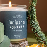 Two Juniper & Cypress soy candles surrounded by pine and juniper accents — clean, woodsy winter fragrance handcrafted by Wax Crescent for cozy seasonal ambiance.