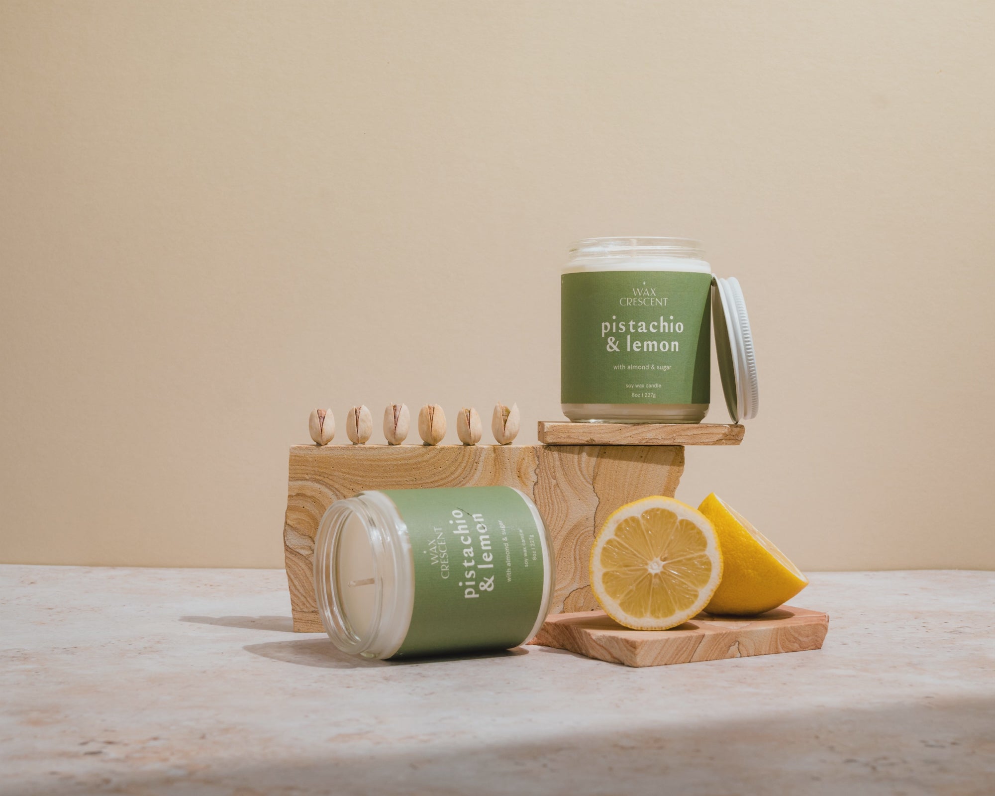 A hand gently lighting a Pistachio & Lemon soy wax candle, surrounded by fresh lemons and pistachios. The flickering flame illuminates the natural ingredients, creating a cozy and inviting atmosphere