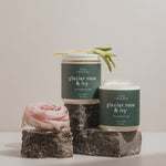 Luxury soy wax candles labeled "Glacier Rose & Ivy," adorned with fresh sage and a rose, set against a neutral background with textured stone props.