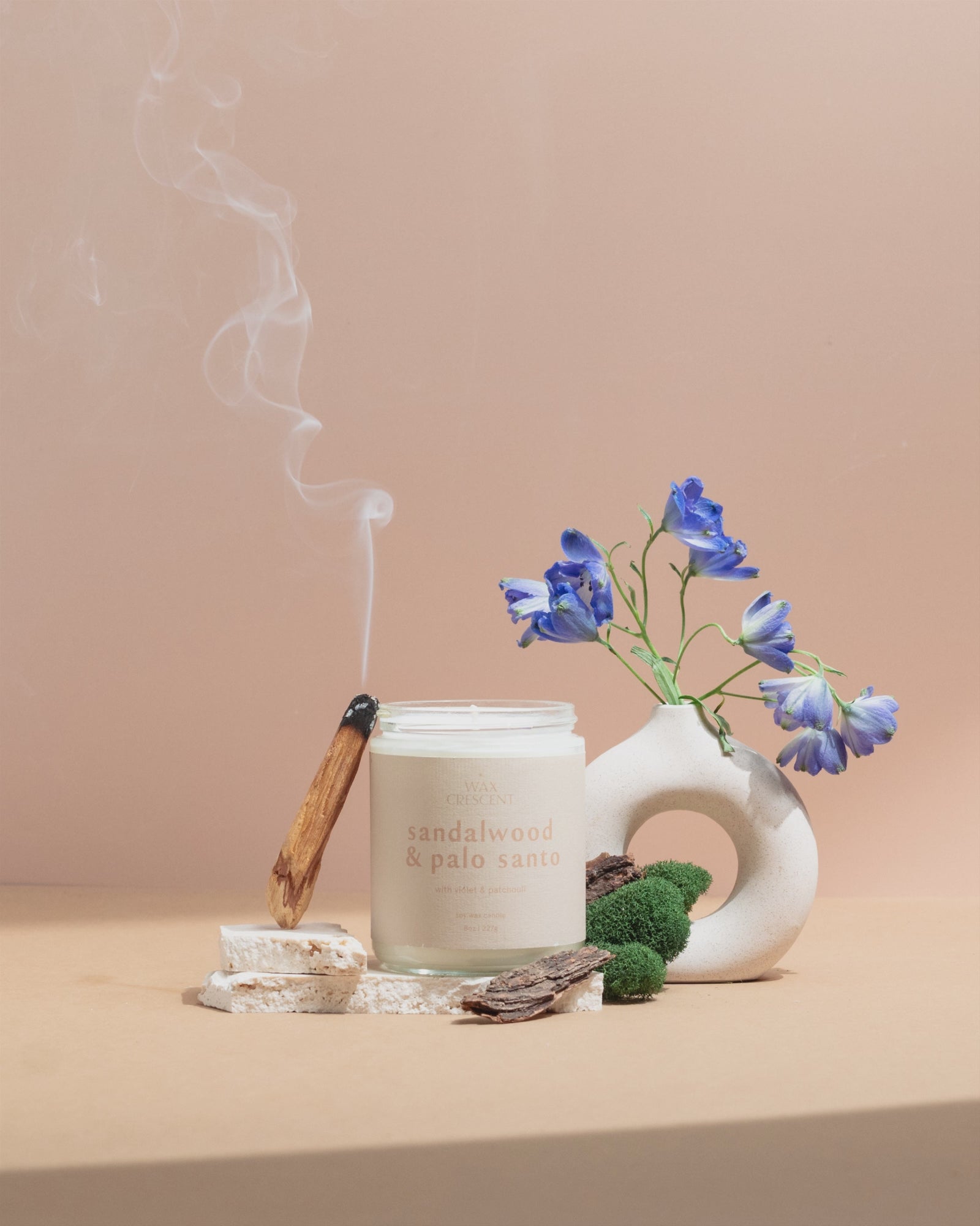 Wax Crescent Sandalwood & Palo Santo luxury soy candle surrounded by natural decor, including palo santo wood, moss, bark, and blue delphinium flowers, styled against a minimalist beige background. Perfect for creating a calming and meditative ambiance with video of palo santo stick smoking 