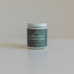 White Birch & Eucalyptus luxury soy wax candle with a white lid, featuring a sleek design and elegant label, placed on a light background with eucalyptus leaves and pinecones, embodying a fresh, forest-inspired aroma.