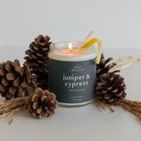 Juniper & Cypress candle surrounded by pinecones and topped with a lemon peel, highlighting its fresh, forest-inspired scent and vibrant citrus notes