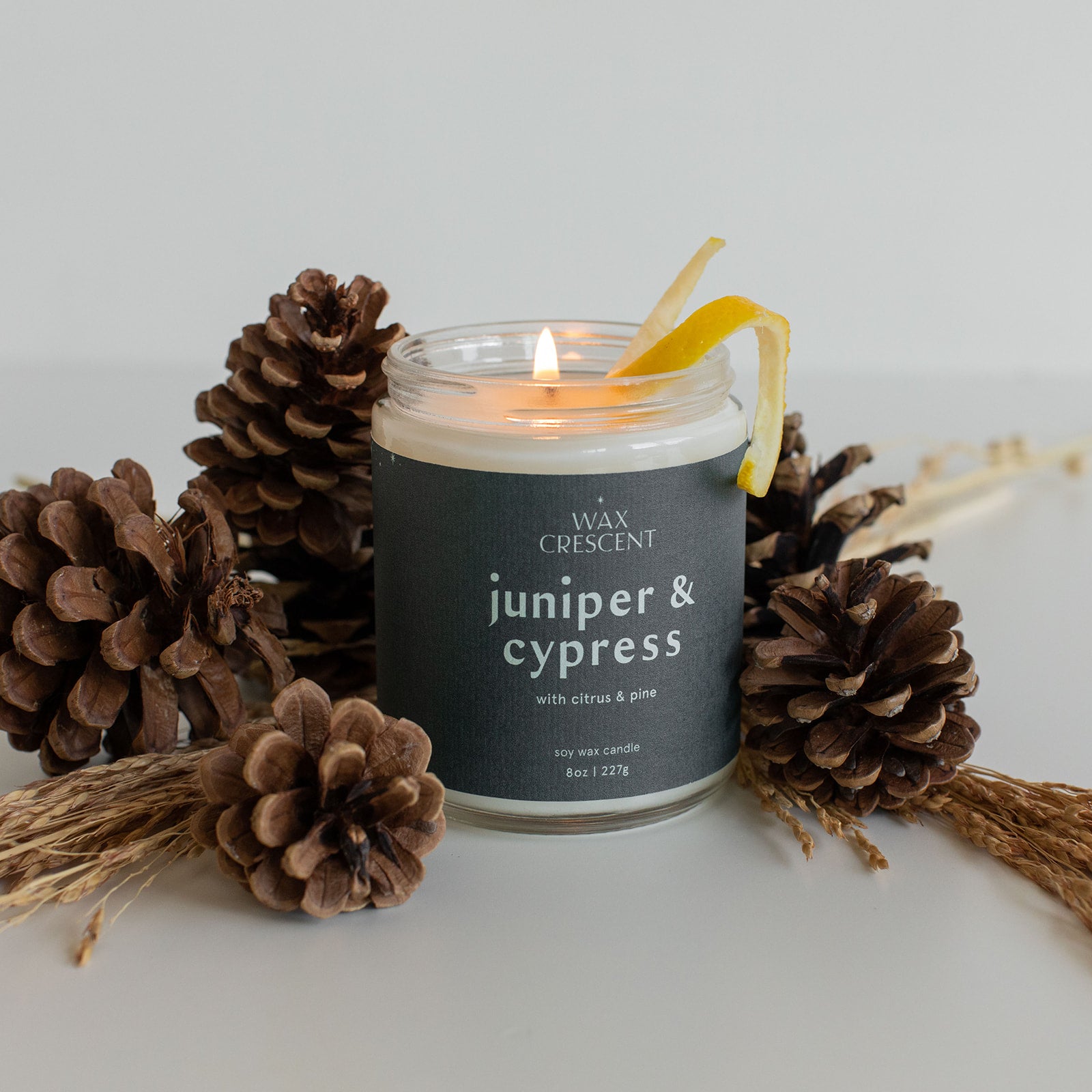 Juniper & Cypress candle surrounded by pinecones and topped with a lemon peel, highlighting its fresh, forest-inspired scent and vibrant citrus notes
