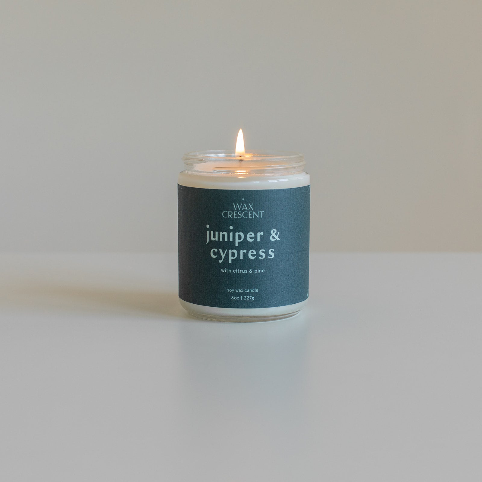 Lit Juniper & Cypress candle with a deep blue label on a black background, showcasing its inviting glow and natural forest-inspired fragrance.