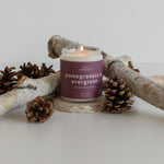Pomegranate & Evergreen candle burning, surrounded by pine cones and branches, showcasing a festive and nature-inspired scent.