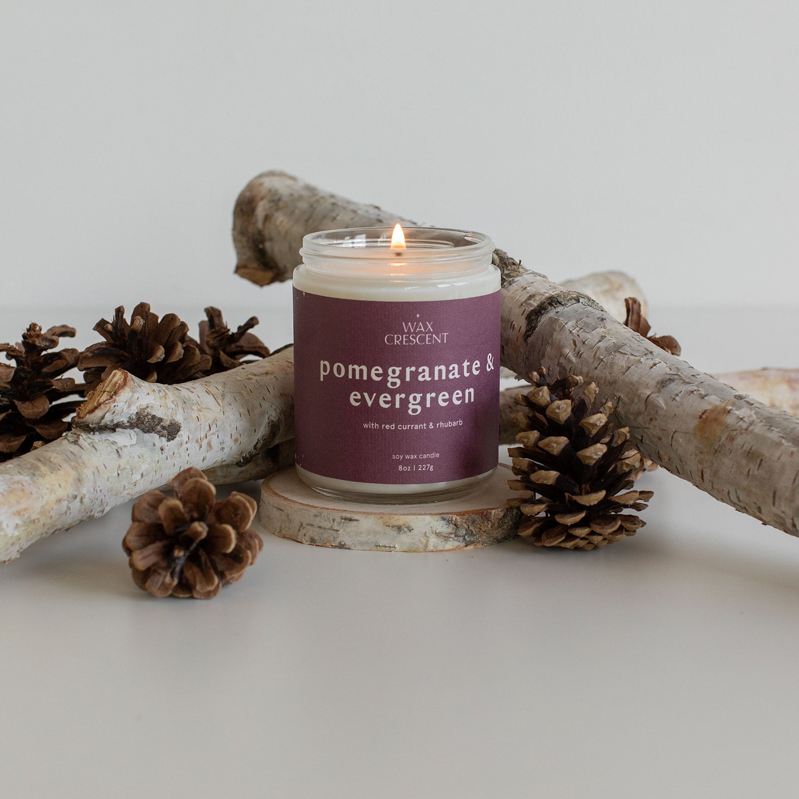 Pomegranate & Evergreen candle burning, surrounded by pine cones and branches, showcasing a festive and nature-inspired scent.