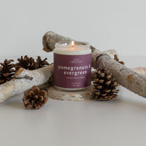 Pomegranate & Evergreen candle burning, surrounded by pine cones and branches, showcasing a festive and nature-inspired scent.