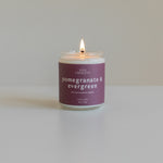 Pomegranate & Evergreen soy candle lit on a plain background, featuring a berry-colored label, hand-poured in Colorado for a vibrant holiday fragrance.