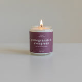 Pomegranate & Evergreen soy candle lit on a plain background, featuring a berry-colored label, hand-poured in Colorado for a vibrant holiday fragrance.