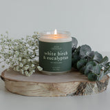 White Birch & Eucalyptus luxury soy wax candle displayed on a wooden surface, highlighting its minimalist design and natural elements, surrounded by eucalyptus leaves and pine branches, evoking a serene forest ambiance.